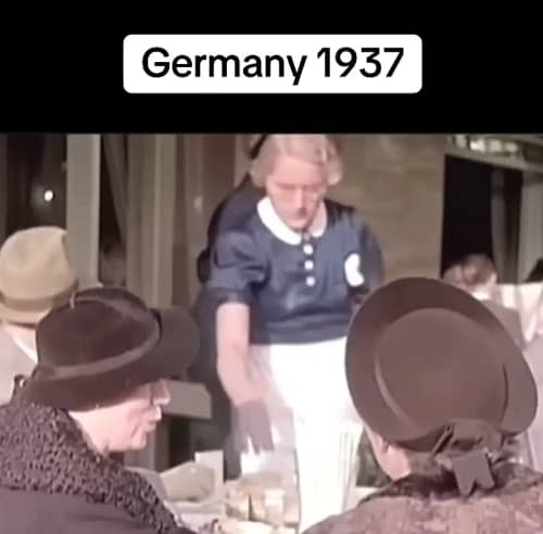 Zwei Frauen mit Hüten sitzen 1937 an einem gedeckten Tisch im Freien, während eine Bedienung Kaffee serviert. Die Frau links im Bild spricht einen Satz, der per Lippenlesen analysiert wurde.